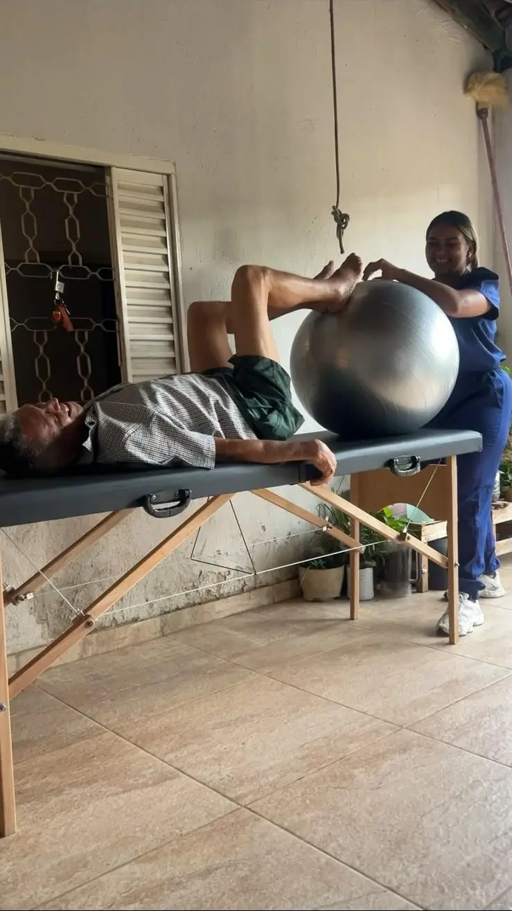 Atendimento domiciliar com bola suíça, paciente deitado e fisioterapeuta sorrindo